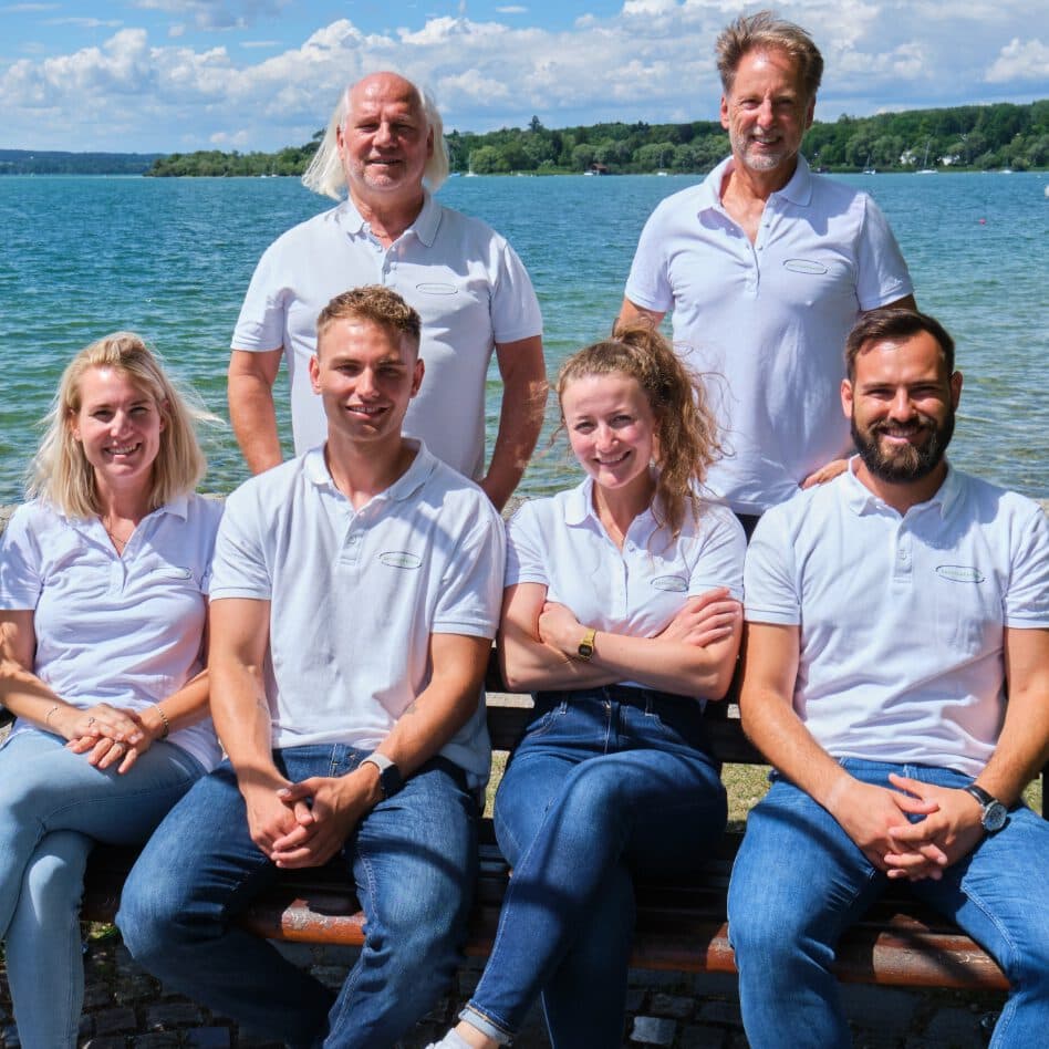Man sieht das ServiceFactum-Team, bestehend aus 6 Personen. Die beiden Geschäftsführer stehen hinter den vier Personen, die auf einer Bank sitzen. Im nahen Hintergrund sieht man den Ammersee.