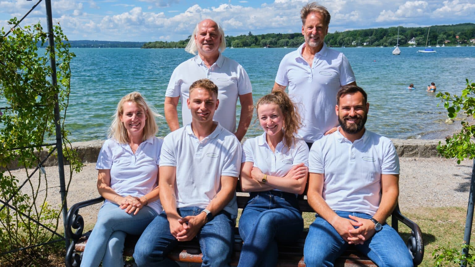 Man sieht das ServiceFactum-Team, bestehend aus 6 Personen. Die beiden Geschäftsführer stehen hinter den vier Personen, die auf einer Bank sitzen. Im nahen Hintergrund sieht man den Ammersee.