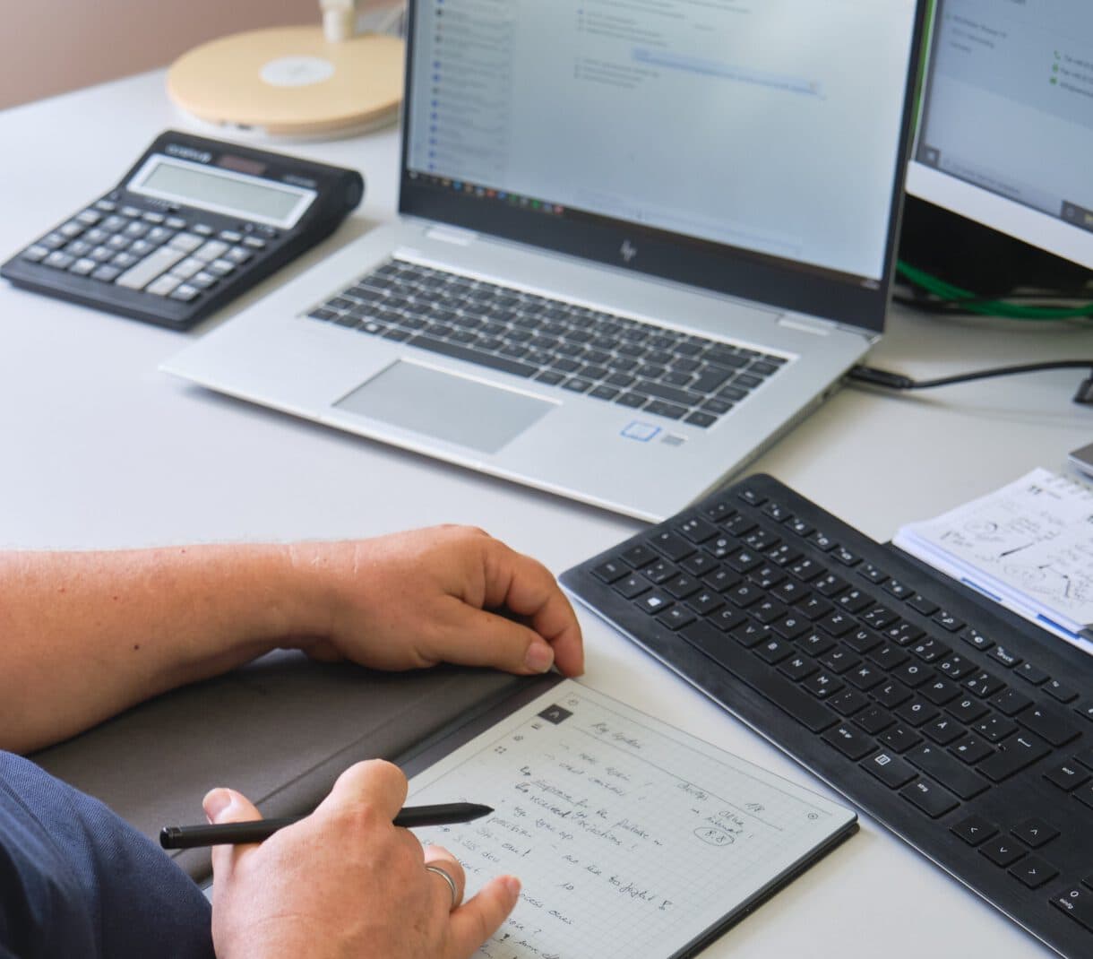Zwei Hände liegen auf einem digitalen Notizbuch auf dem Schreibtisch. Die rechte Hand hält einen Stift. Im Hintergrund sieht man einen Taschenrechner und einen aufgeklappten Laptop.