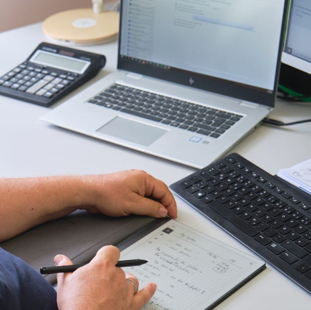 Zwei Hände liegen auf einem digitalen Notizbuch auf dem Schreibtisch. Die rechte Hand hält einen Stift. Im Hintergrund sieht man einen Taschenrechner und einen aufgeklappten Laptop.