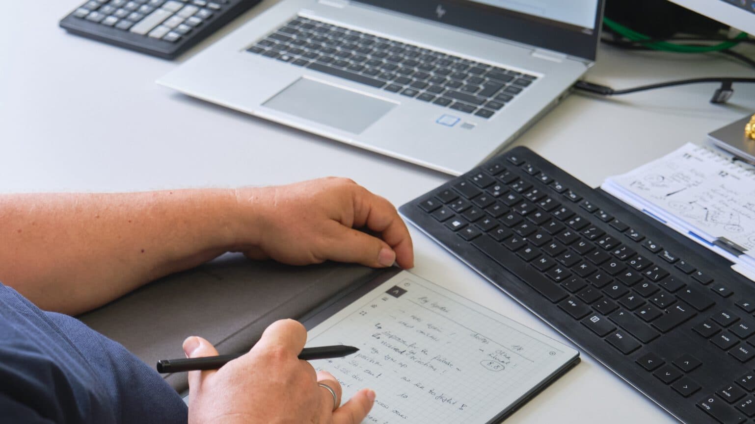 Zwei Hände liegen auf einem digitalen Notizbuch auf dem Schreibtisch. Die rechte Hand hält einen Stift. Im Hintergrund sieht man einen Taschenrechner und einen aufgeklappten Laptop.