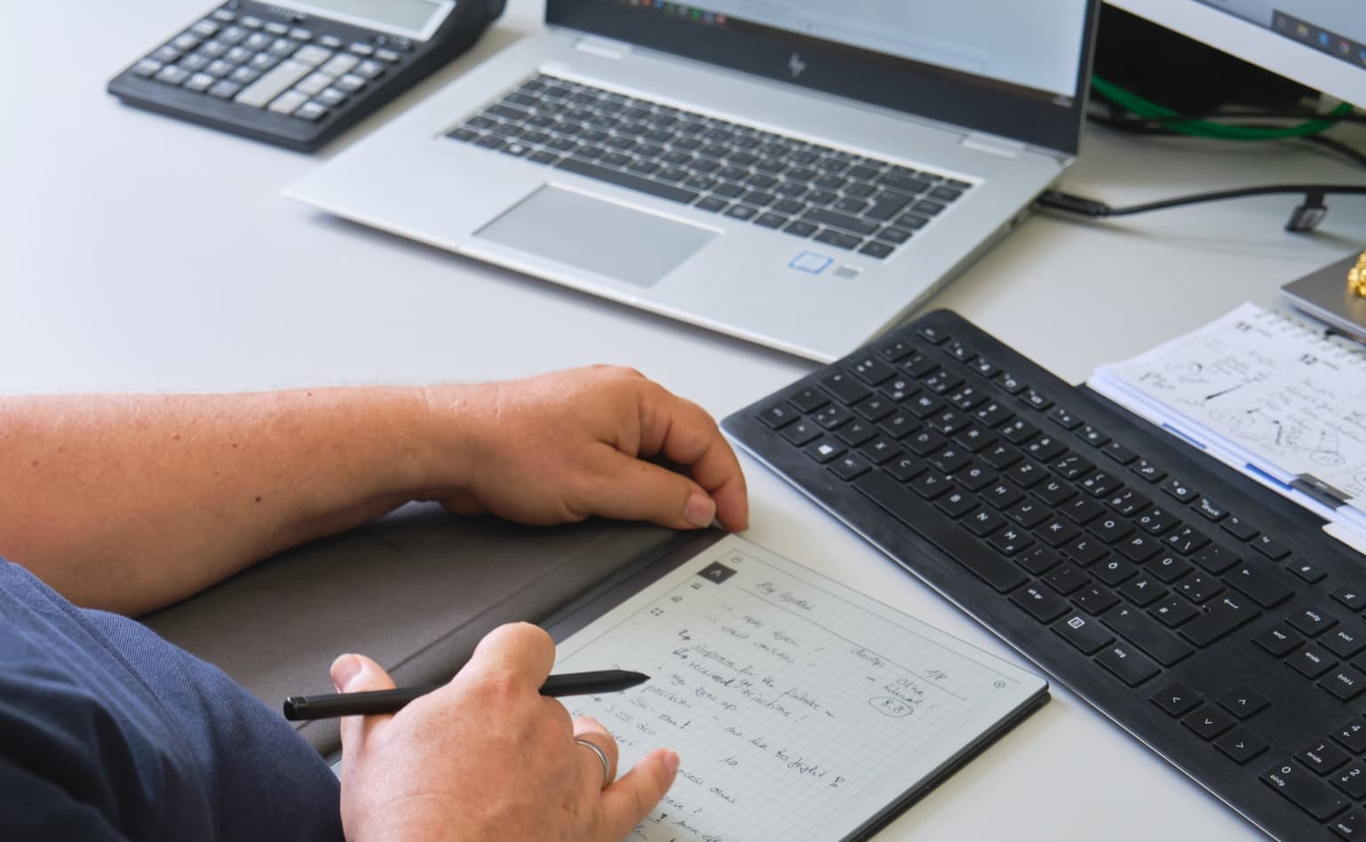 Zwei Hände liegen auf einem digitalen Notizbuch auf dem Schreibtisch. Die rechte Hand hält einen Stift. Im Hintergrund sieht man einen Taschenrechner und einen aufgeklappten Laptop.