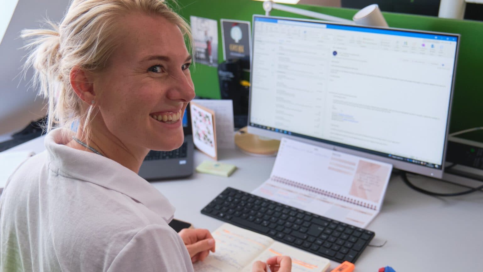 Frau, die lachend an ihrem Schreibtisch sitzt und nach hinten umgekehrt ihren Blick richtet. Sie hält ihren Stift in der Hand und ihre Hände liegen auf ihrem Notizblock. Im Hintergrund sieht man einen Bildschirm, welcher an ist.