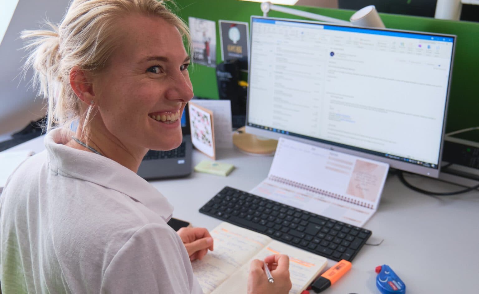 Frau, die lachend an ihrem Schreibtisch sitzt und nach hinten umgekehrt ihren Blick richtet. Sie hält ihren Stift in der Hand und ihre Hände liegen auf ihrem Notizblock. Im Hintergrund sieht man einen Bildschirm, welcher an ist.