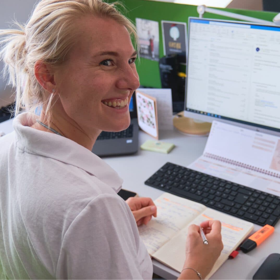 Frau, die lachend an ihrem Schreibtisch sitzt und nach hinten umgekehrt ihren Blick richtet. Sie hält ihren Stift in der Hand und ihre Hände liegen auf ihrem Notizblock. Im Hintergrund sieht man einen Bildschirm, welcher an ist.