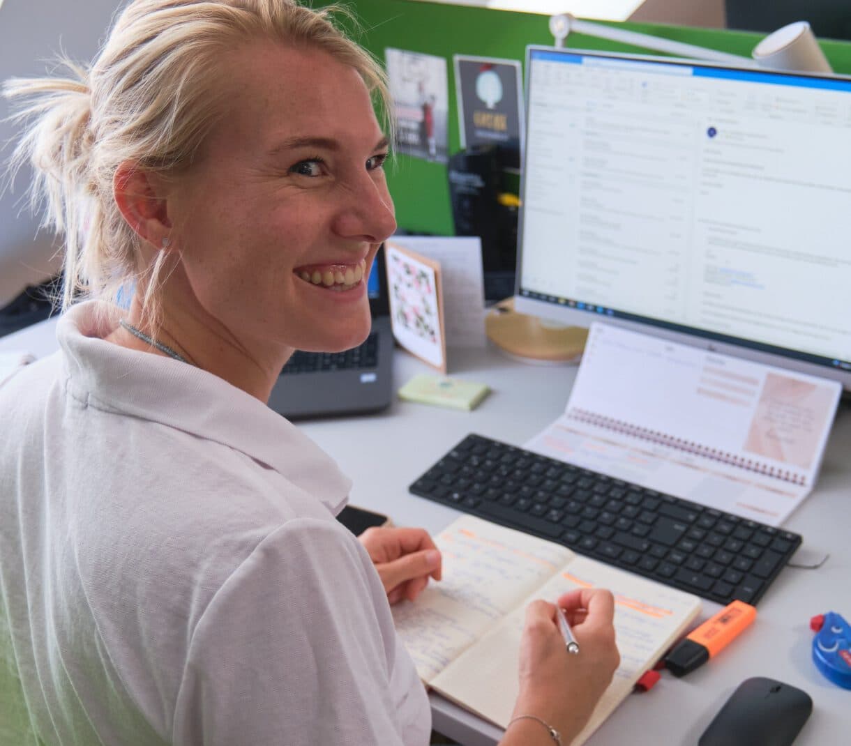 Frau, die lachend an ihrem Schreibtisch sitzt und nach hinten umgekehrt ihren Blick richtet. Sie hält ihren Stift in der Hand und ihre Hände liegen auf ihrem Notizblock. Im Hintergrund sieht man einen Bildschirm, welcher an ist.
