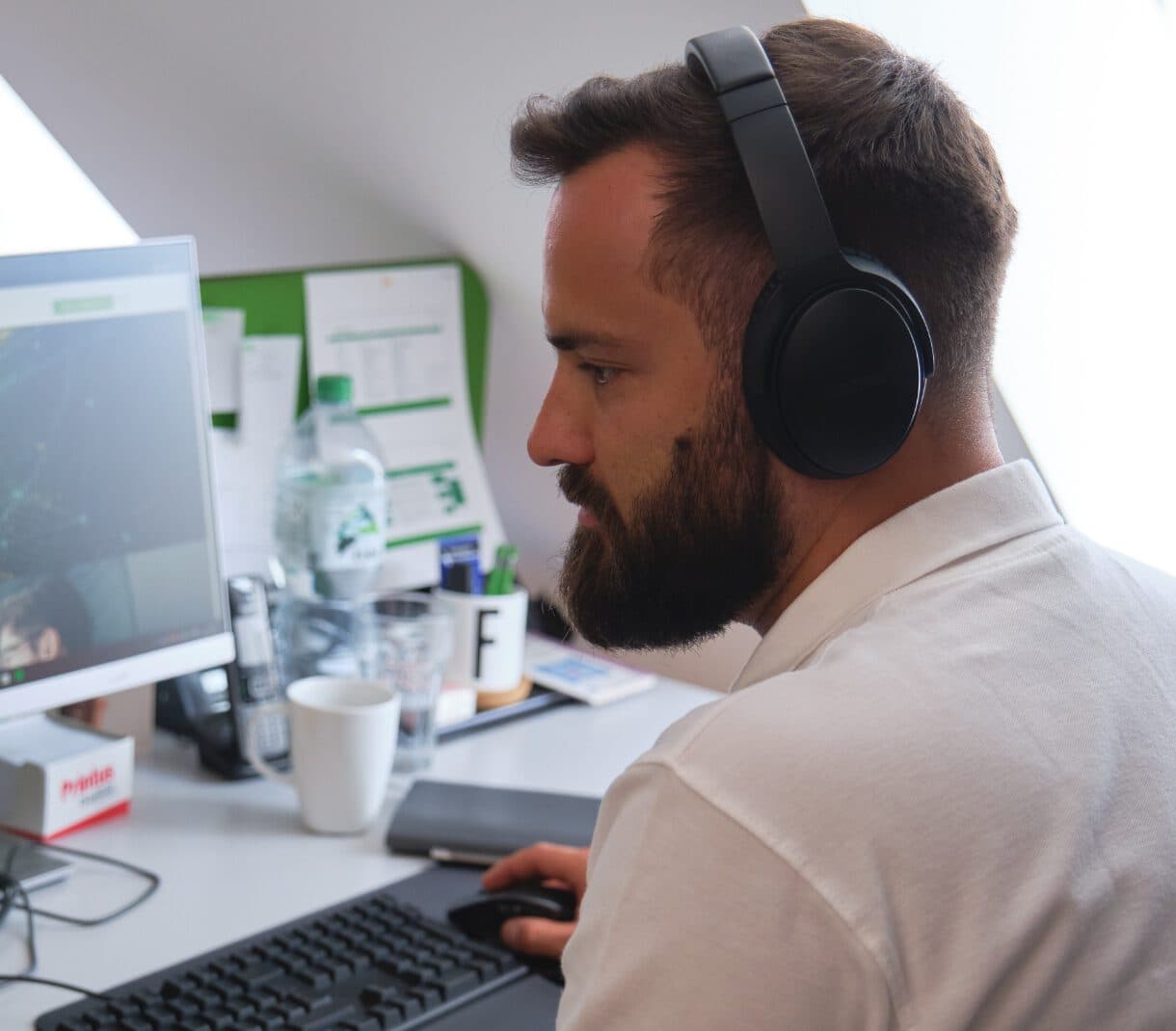 Mann mit Bart und Kopfhörer, der am Schreibtisch sitzt und seine rechte Hand hält die Maus vom Computer. Im Hintergrund sieht man einen Bildschirm, Wasserflasche, Kaffeebecher und ein Notizbuch.