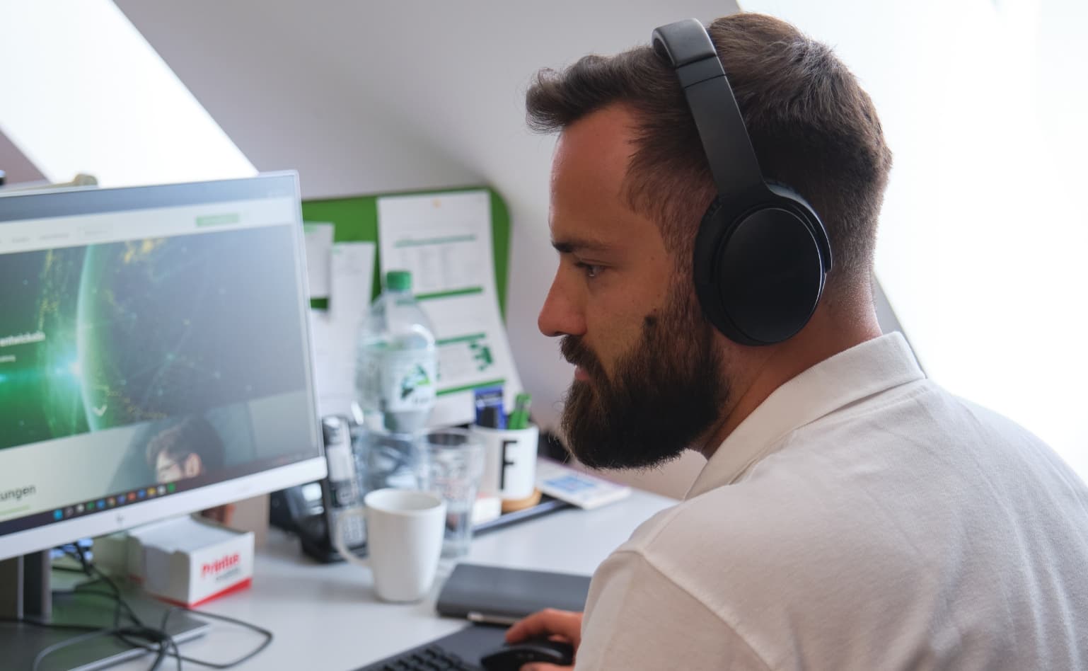 Mann mit Bart und Kopfhörer, der am Schreibtisch sitzt und seine rechte Hand hält die Maus vom Computer. Im Hintergrund sieht man einen Bildschirm, Wasserflasche, Kaffeebecher und ein Notizbuch.