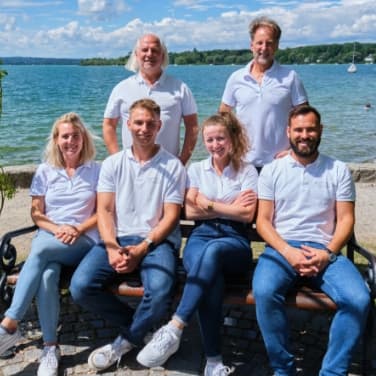 Man sieht das ServiceFactum-Team, bestehend aus 6 Personen. Die beiden Geschäftsführer stehen hinter den vier Personen, die auf einer Bank sitzen. Im nahen Hintergrund sieht man den Ammersee.