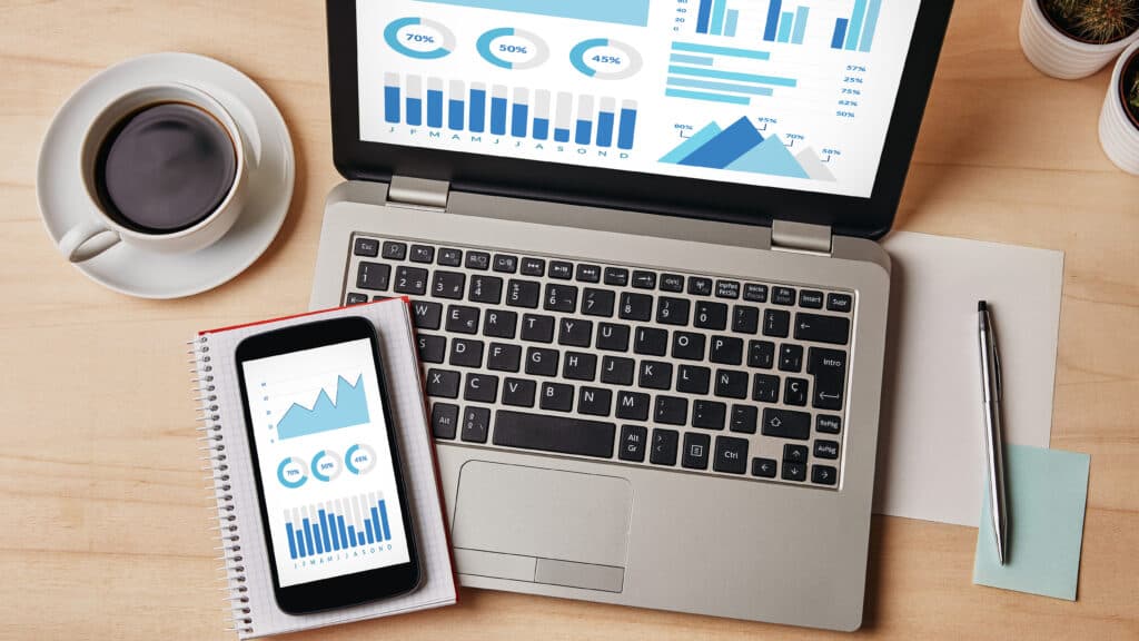 Grafiken und Charts auf einem Handy und Laptop. Man sieht auch eine volle Kaffeetasse und Notizen und einen Stift.