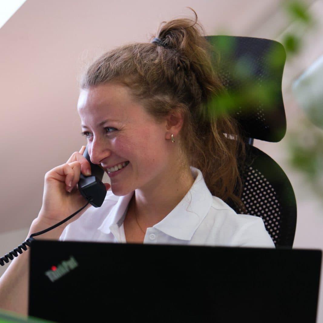 Frau, die lachend ein Telefon in ihrer rechten Hand hält und die ihren Blick nach links gerichtet ist. Im Vordergrund sieht man verschwommen eine Pflanze und einen geöffneten Laptop.