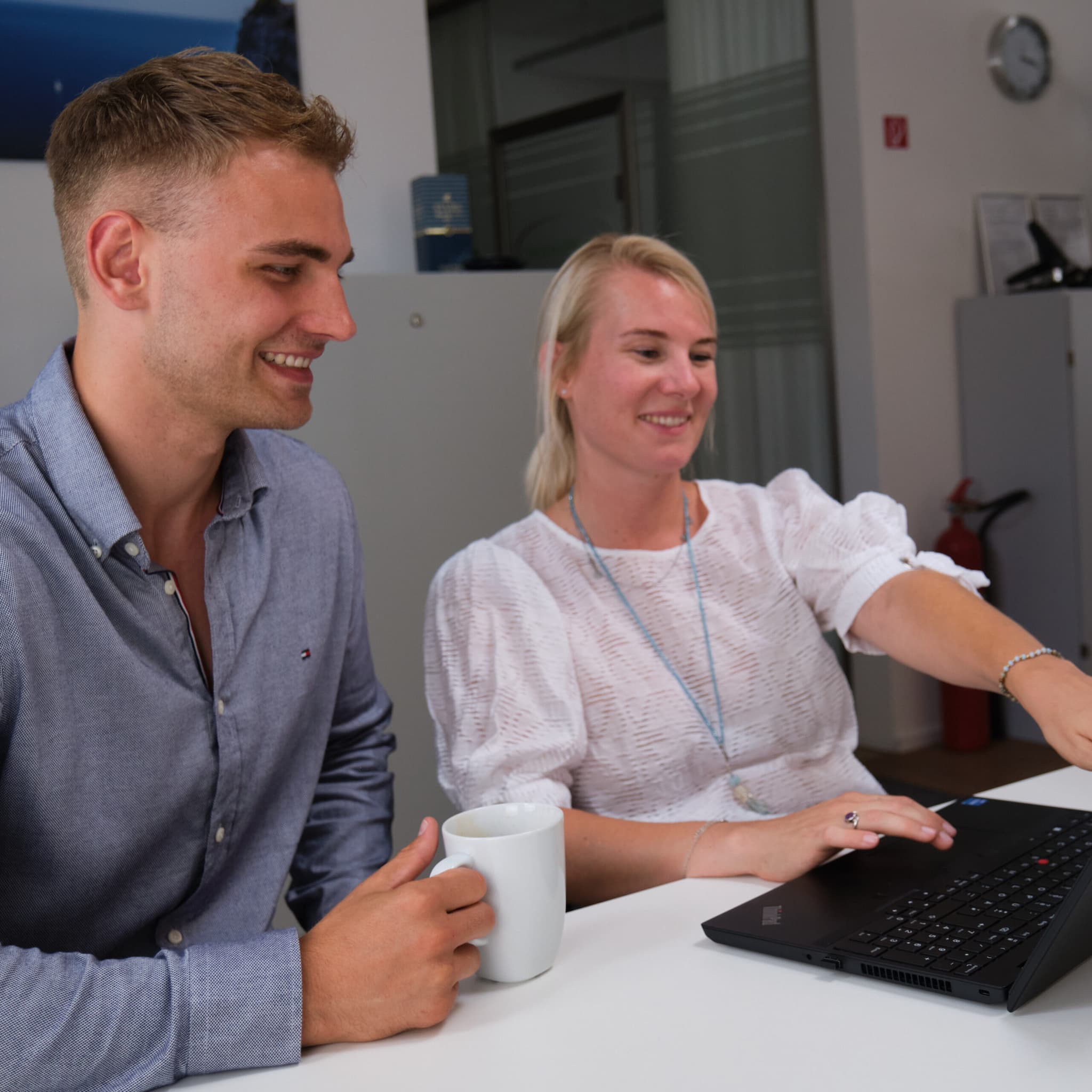 Zwei Mitarbeitende von ServiceFactum, die lachend auf einen Laptop schauen. Beide sitzen an einem Tisch. Die Frau zeigt auf den Bildschirm. Der Mann hat ein Kaffeeglas in der rechten Hand.