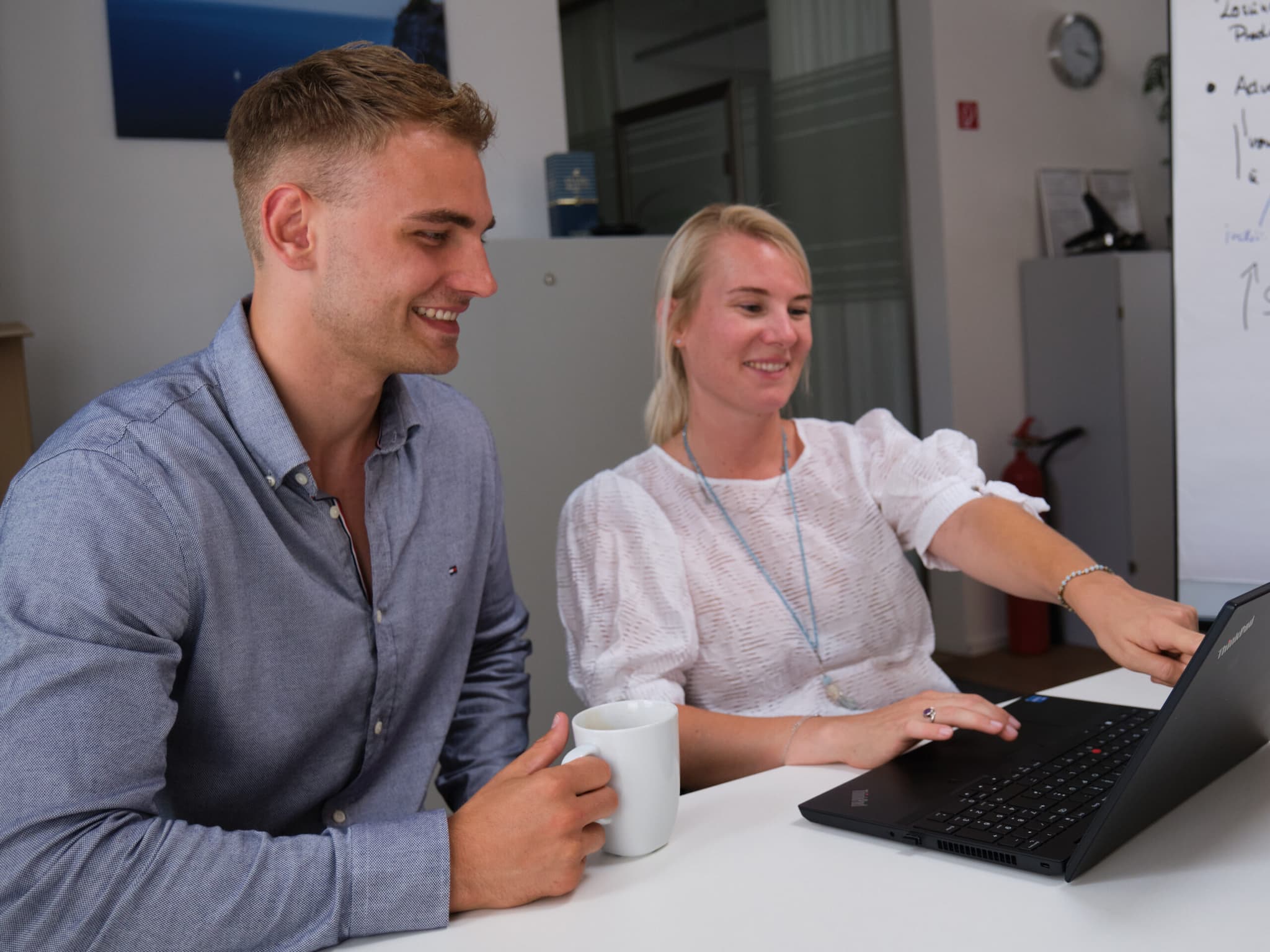 Zwei Mitarbeitende von ServiceFactum, die lachend auf einen Laptop schauen. Beide sitzen an einem Tisch. Die Frau zeigt auf den Bildschirm. Der Mann hat ein Kaffeeglas in der rechten Hand.